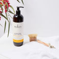 Sukin hydrating body lotion bottle with a wooden brush on a white surface with flowers in the background